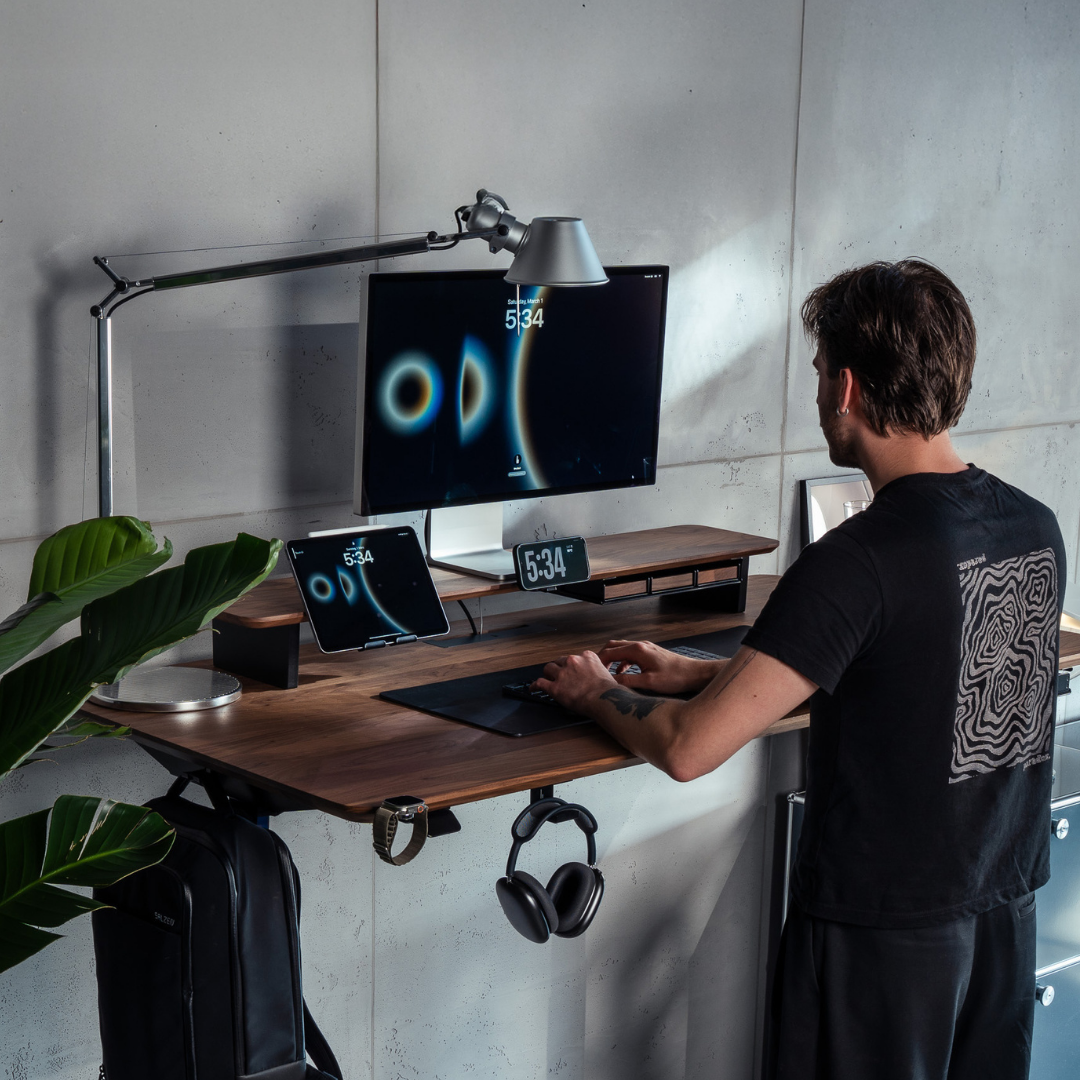 Ein Mann steht an einem höhenverstellbaren BALOLO Desk Cockpit aus Holz mit schwarzem Mteallgestell. Auf dem Schreibtisch befinden sich Monitor, Tablet und Smartphone in passenden BALOLO Add-Ons, die an dem Cockpit montiert sind.
