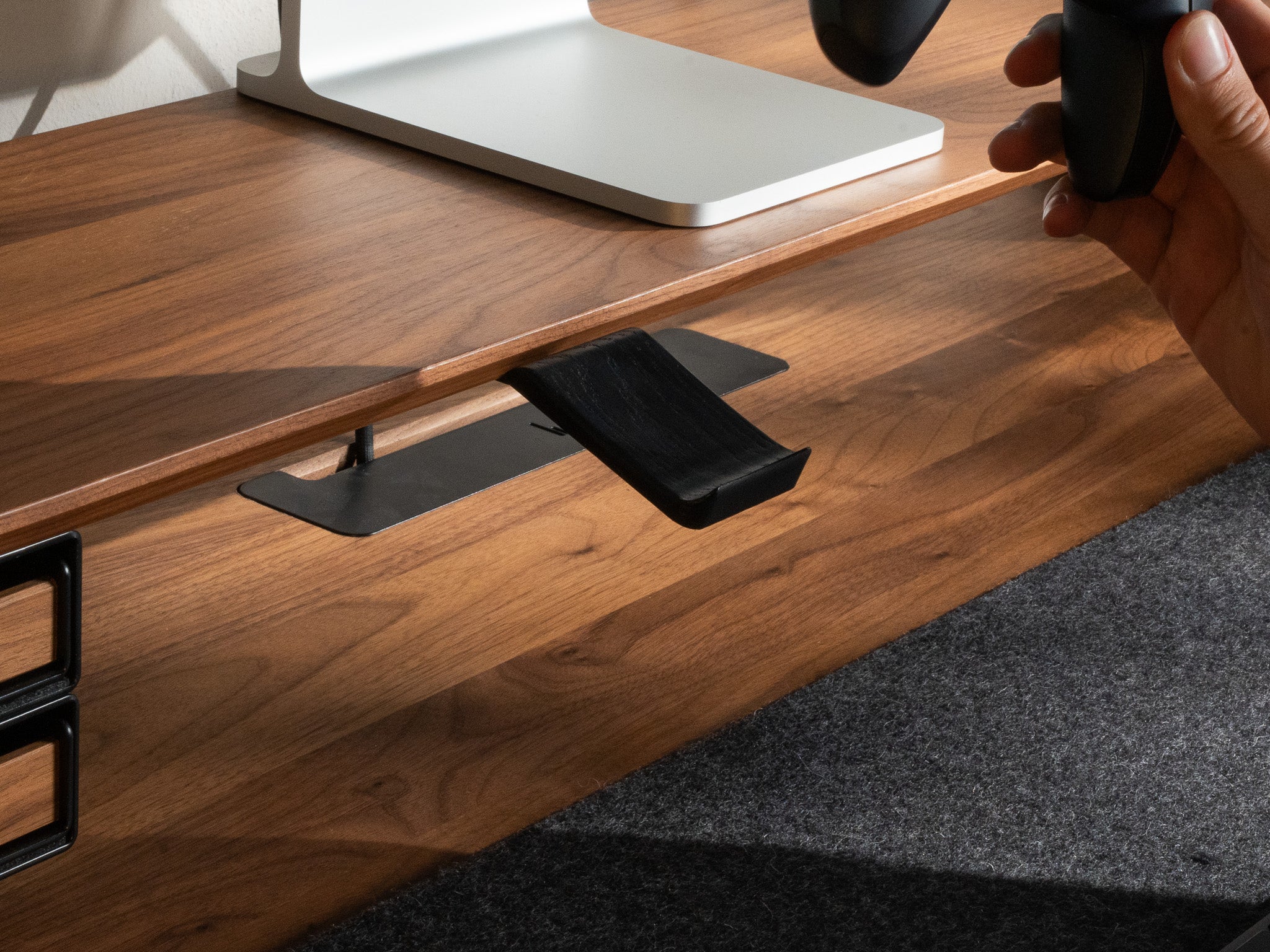 Detail-Aufnahme vom BALOLO Controller Halter montiert am Setup Cockpit Monitorständer in Nussbaum. In ästhetischem Office-Setup, Hand nimmt in Bewegung einen PlayStation 5 Controller aus der Controller Halterung.