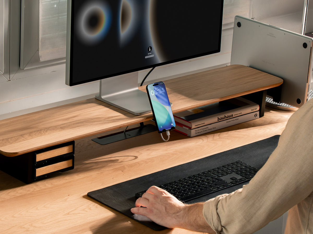 Setup Cockpit aus Eichenholz mit Monitor, Smartphone im Handyhalter, senkrecht stehendem Laptop und Schubladen auf einem aufgeräumten Schreibtisch mit schwarzer Schreibtischunterlage @expand #material_Eiche