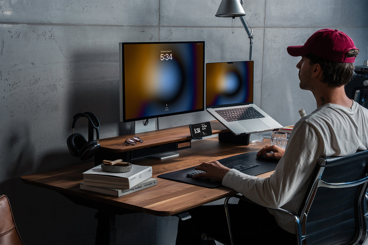 Mann mit roter Kappe sitzt am Schreibtisch mit BALOLO Setup Cockpit aus Nussbaum, Monitor, Laptop und Handyhalter